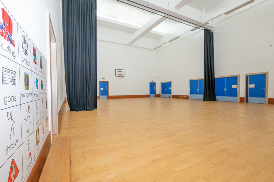 Image of a school sports hall, with high ceiling and a curtain that can divide the room. There is a board on the wall with symbols to aid communication. 